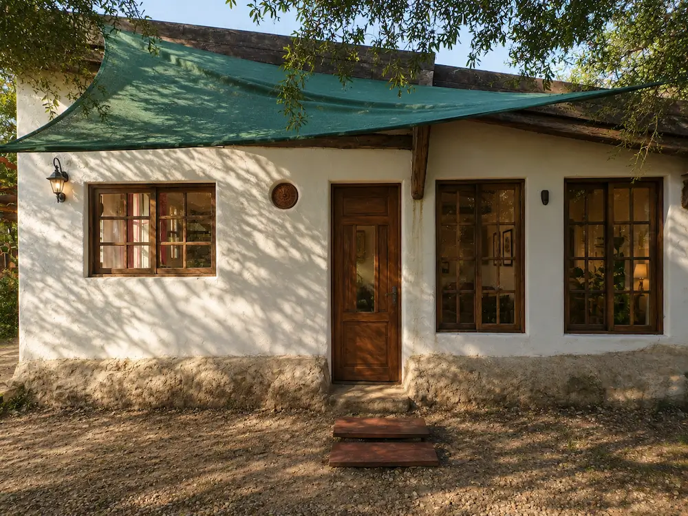 Small rustic house with white walls, wooden door, three windows, and shade sail overhead outdoors.