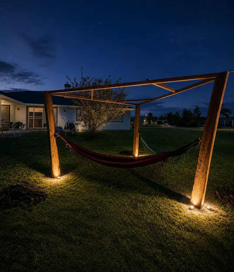 Wooden triangular hammock stand with lights at base, set on grass in a backyard at night.